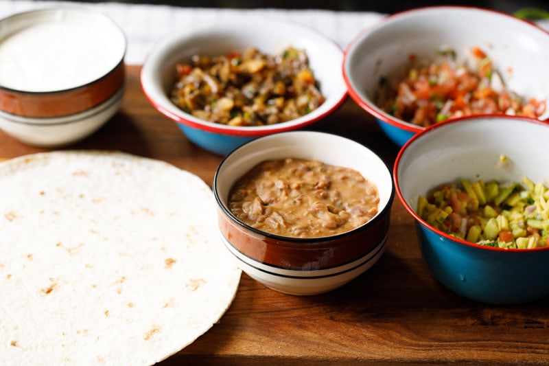 flour tortilla on a wooden surface with bowls filled with vegetarian burrito stuffings like refried beans, guacamole, pico de gallo and fajita style veggies