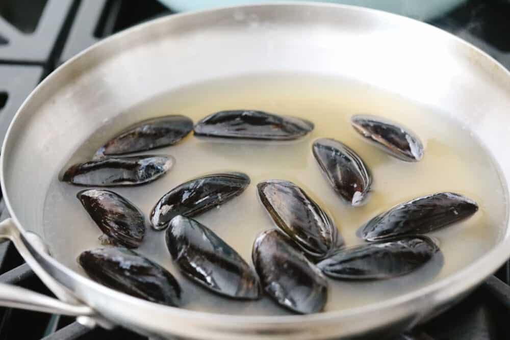 These steamed mussels are easy, elegant, and delicious! They are perfect for any dinner party and you won't be able to stop after just one! Uncooked mussels in a pan with white wine and chicken broth.